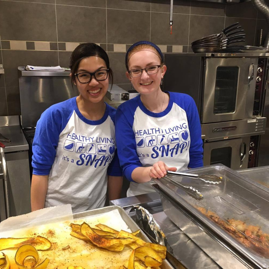 two SNAP volunteers standing behind a cooking station wearing SNAP shirts