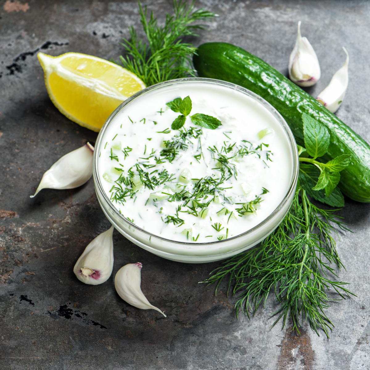 A bowl of Tzatziki with garlic, herbs, lemon and a cucumber around it.